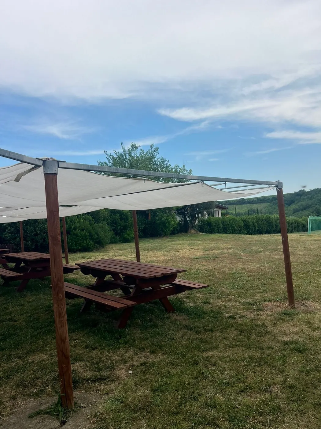 Un gazebo con telo bianco ombreggiante e tavoli di legno su un prato; sullo sfondo siepi e colline coltivate