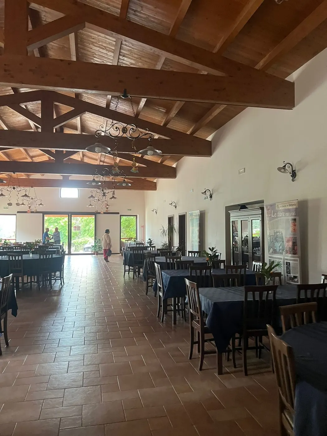 La sala interna, con travi di legno a vista sul soffitto, tavoli rotondi con tovaglie blu, sedie e grandi porte a vetri sul verde